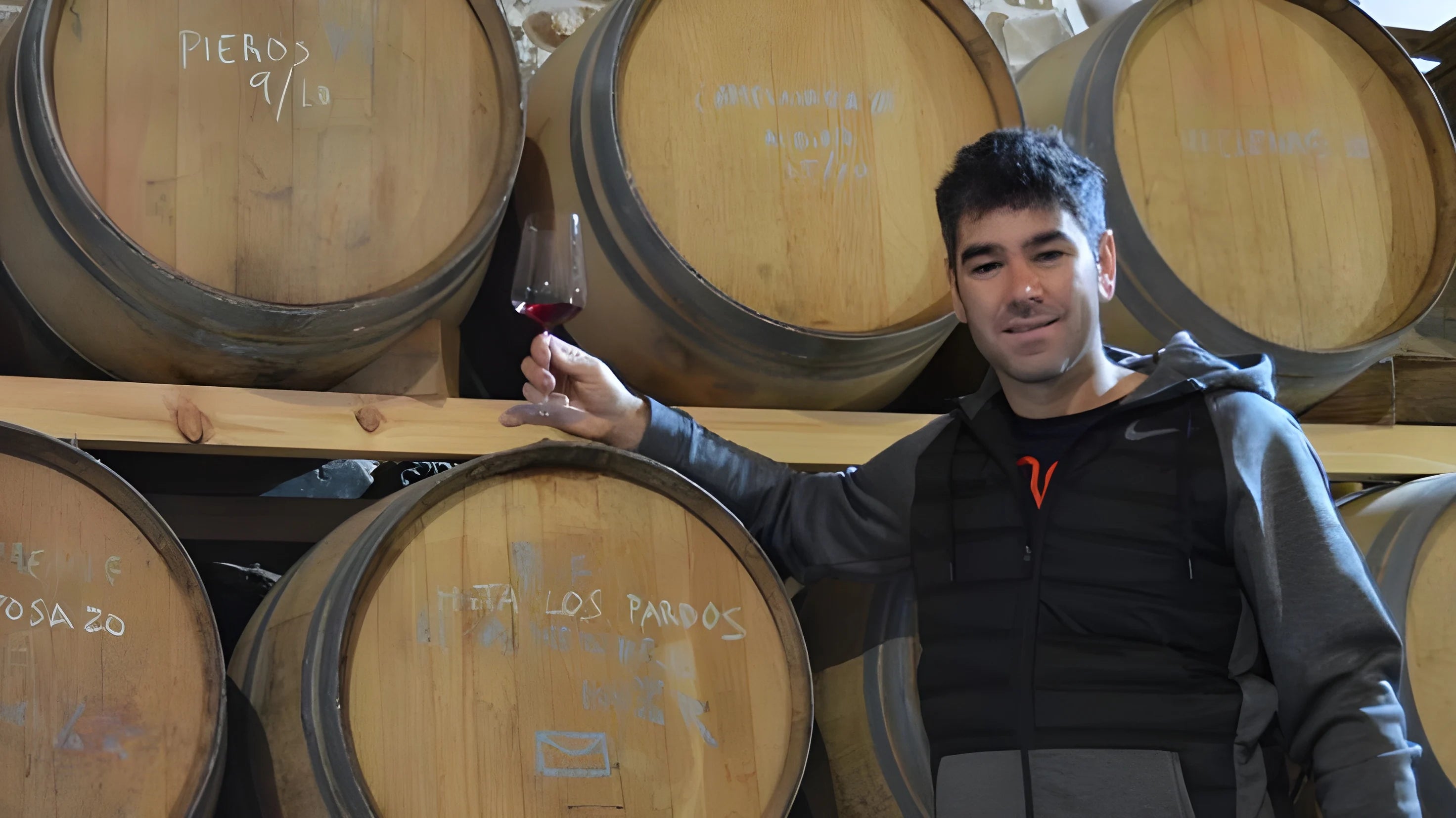 Winemaker César Márquez holding a glass of red wine in his cellar in El Bierzo, Spain, surrounded by oak aging barrels.