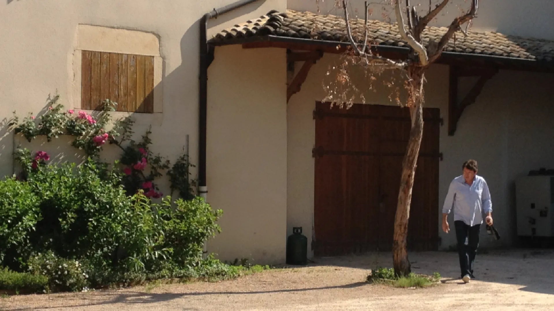 Jean Foillard walking outside a rustic building at his estate, with flowers and a gravel path on a sunny day.