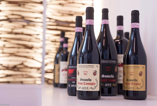 A curated lineup of Arpepe Valtellina Superiore 2001 wines, featuring Grumello Buon Consiglio, Sassella Rocce Rosse and Sassella Regina, displayed on a white surface with a wooden backdrop.
