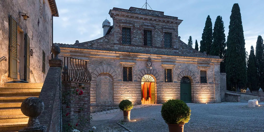 Fèlsina historic stone building at dusk with arched windows and doors, warmly lit entrance, surrounded by cypress trees and glowing potted shrubs.