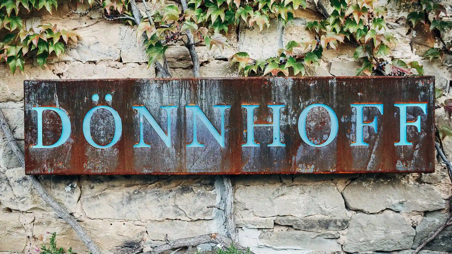 Rustic metal sign reading “DÖNNHOFF” mounted on a stone wall with ivy, marking the entrance to the historic Dönnhoff winery. The aged textures and creeping vines evoke tradition, craftsmanship, and the natural elegance of fine winemaking