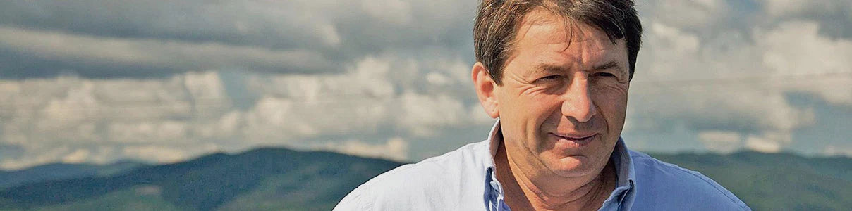 Portrait of Jean Foillard standing outdoors with rolling green hills and a cloudy sky in the background, wearing a light blue shirt and looking toward the camera.
