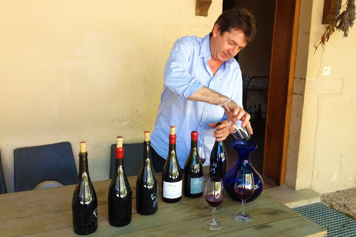 Jean Foillard pouring red wine into a decanter at his estate, with bottles and glasses arranged on a rustic outdoor table
